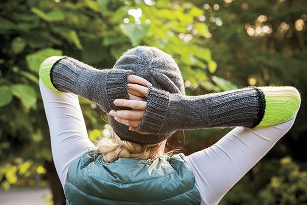 A woman is shown with her back to the camera, standing in a wooded area, modeling a gray knitted hat and knitted arm warmers in gray and neon yellow. The Brightwood Hat & Mitts pattern, a design by Tian Connaughton for Knit Picks.