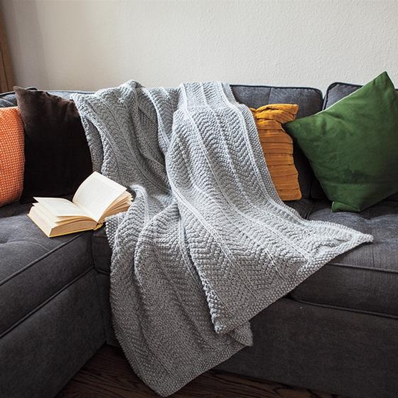 A gray knitted blanket draped on a couch. The Abrigado blanket pattern: Made in bulky yarn with an arrow pattern pleasing to the eye, you'll want one for every room in your home.