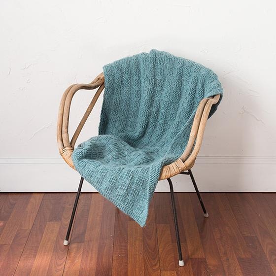 A green knitted blanket with a basketweave pattern is draped on a wicker chair