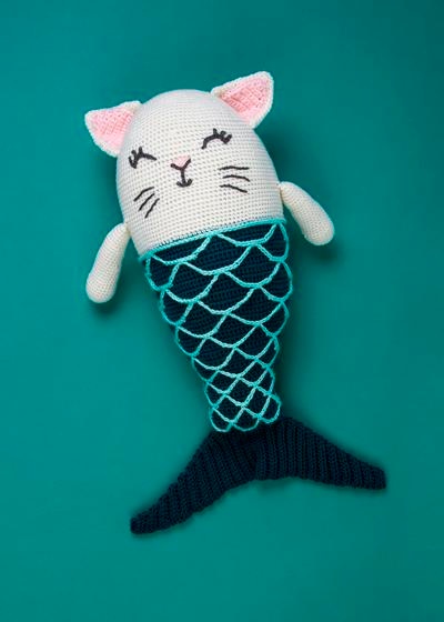 A crocheted toy with a white cat body on top and a dark green mermaid tail on bottom.