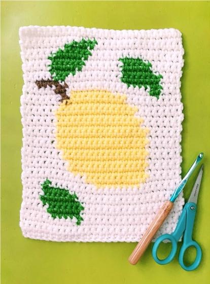 An image of a crochet block with a lemon design on it. A crochet hook and scissors.