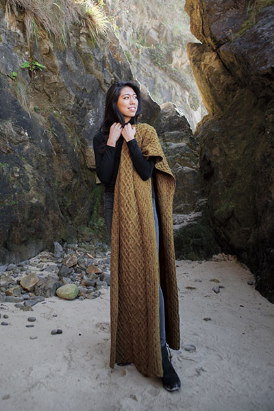 A woman wearing a long cabled rectangular shawl.  The shawl is draped over her shoulder and falls to her feet. 